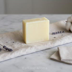 May include: A light yellow rectangular soap bar sits on a beige linen cloth, with sprigs of lavender and a cotton flower nearby. The text "Esoteric Aroma" is visible at the bottom of the image.