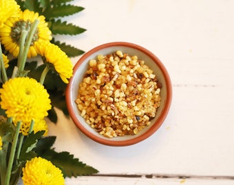 May include: A small brown bowl filled with golden brown resin incense. The bowl is sitting on a white wooden surface. There are yellow flowers in the background.