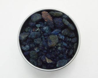 May include: A small metal container filled with dark blue and black mineral chips.