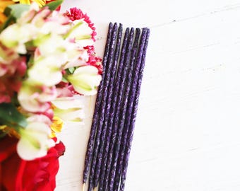 May include: A collection of purple incense sticks with a textured design, placed diagonally on a white wooden surface. A vibrant floral arrangement featuring red, yellow, and white flowers is in the background.
