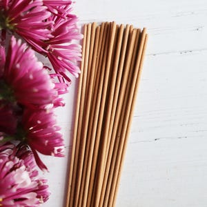 May include: A bunch of pink flowers with a bundle of brown wooden sticks on a white wooden surface.