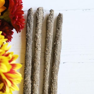 May include: Four cylindrical incense sticks with a rough, textured, grey-brown surface arranged vertically next to a cluster of vibrant yellow and deep red chrysanthemum flowers on a white surface.