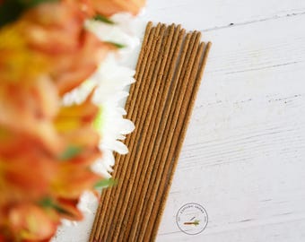 May include: A bundle of brown incense sticks arranged diagonally on a white wooden surface. The incense is next to a blurred arrangement of orange and white flowers. A small logo is in the bottom right corner.