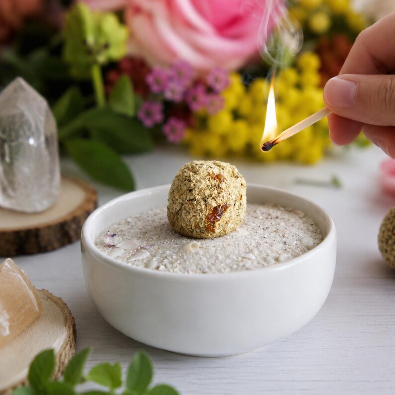 Incense ball in sand in a white bowl with crystals and flowers nearby