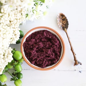 May include: A small bowl filled with a deep purple powder, likely acai powder, sits on a white surface.  A decorative spoon with a crystal handle is also on the surface.  White flowers and green leaves are in the background.
