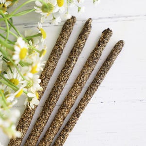 May include: Five brown incense sticks with a textured surface are arranged diagonally on a white wooden surface. Chamomile flowers with white petals and yellow centers are in the upper left corner. The incense sticks have a speckled appearance.