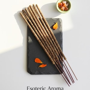 May include: A collection of brown incense sticks arranged on a black slate, with orange flower petals and a small green bowl filled with orange stones. The text "Esoteric Aroma" is at the bottom of the image.