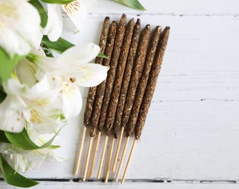May include: A bundle of incense sticks with a dark brown, textured appearance, resting on a white wooden surface. White lilies with green leaves are arranged to the left of the incense. The incense sticks are on thin wooden sticks.