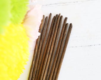 May include: A collection of brown incense sticks, positioned diagonally against a white wooden backdrop. The incense sticks are long and thin, with a slightly rough texture. Soft yellow, pink, and green accents are visible in the upper left corner.
