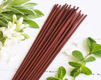 May include: A collection of long, slender, dark reddish-brown wooden sticks arranged in a diagonal cluster. The sticks have a smooth, matte finish and are positioned alongside fresh green sage leaves, white flowers, and a sprig of green mint.