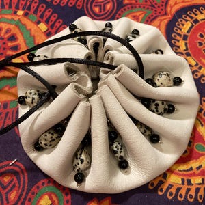 May include: A white leather pouch with a drawstring closure. The pouch is adorned with black and white speckled beads and black beads. The background features a colorful patterned fabric.