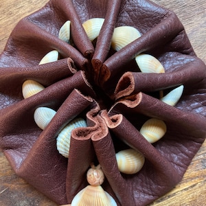 May include: A dark brown leather pouch, gathered at the top, adorned with seashells. The pouch is decorated with a seashell pendant and a thin, light brown cord. The seashells are white and tan.