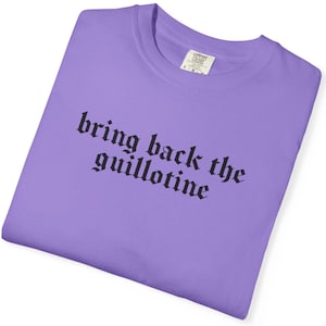 May include: A lilac-colored crew neck t-shirt with the phrase "bring back the guillotine" printed in black gothic font. The shirt has a tag in the upper right corner. The fabric appears soft and comfortable.