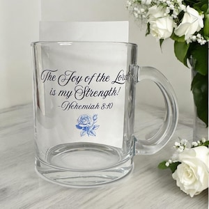 May include: A clear glass mug featuring the black printed text The Joy of the Lord is my Strength! -Nehemiah 8:10 and a small blue rose illustration. The mug has a sturdy handle and a smooth, transparent finish, showcasing the design clearly.