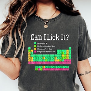 Puede incluir: Camiseta gris oscuro con la frase "Can I Lick It?" y un diseño colorido de la tabla periódica. La camiseta incluye una leyenda con frases como "Claro, hazlo" y "Quizás no sea la mejor idea."