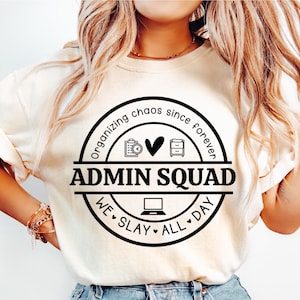 May include: A cream-coloured t-shirt featuring a black circular graphic with the text "ADMIN SQUAD", "Organising chaos since forever", and "We slay all day". The shirt is worn with light blue denim jeans.