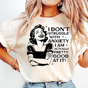 Könnte beinhalten: Cremefarbenes T-Shirt mit dem Text "I DON'T STRUGGLE WITH ANXIETY I AM ACTUALLY PRETTY GOOD AT IT!" und einer Schwarz-Weiß-Illustration einer lachenden Frau. Das Shirt wird mit hellblauen Jeans kombiniert.