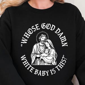 May include: Black sweatshirt with a white graphic. The graphic is a circular design with an illustration of a family and the text "WHOSE GOD DAMN WHITE BABY IS THIS?" in a bold, sans-serif font. The sweatshirt is a casual style.