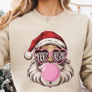 Może przedstawiać: Beżowa bluza z grafiką Świętego Mikołaja w okularach w kształcie serca z napisem "MERRY CHRISTMAS" i różową bańką z gumy do żucia. Bluza jest połączona z czerwono-białymi spodniami od piżamy w kratę. Widoczny jest również biały kubek z grafiką Świętego Mikołaja.