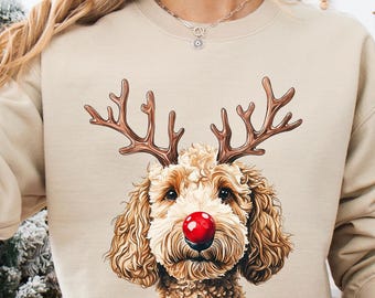 May include: Beige sweatshirt featuring a dog with reindeer antlers and a red nose. The dog graphic is centered on the front of the sweatshirt. The person is wearing red and white plaid pajama pants and holding a white mug with a Santa Claus graphic.