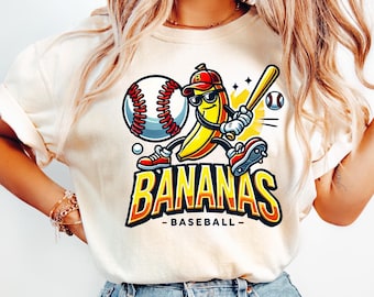 May include: Cream-colored t-shirt featuring a cartoon banana playing baseball. The banana wears a red cap, sunglasses, and cleats, holding a bat. The shirt has the words "BANANAS - BASEBALL-" in orange and yellow.