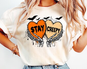 May include: Cream-coloured t-shirt with a Halloween graphic. The design features two heart-shaped elements, one orange and one yellow, with the words "STAY CREEPY". Skeleton hands hold the hearts, and bats are above.
