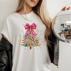 Pode incluir: Camiseta branca com um design colorido de árvore de Natal e um laço rosa. A árvore é decorada com luzes e bolas. Uma bola de discoteca prateada é segurada no fundo. A camiseta é um artigo de vestuário festivo.
