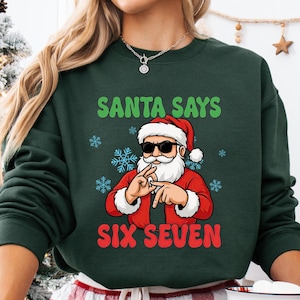 May include: Dark green sweatshirt with the text "SANTA SAYS SIX SEVEN" and a graphic of Santa Claus wearing sunglasses. The person in the image is wearing red and white plaid pajama pants and holding a mug with a Santa Claus graphic.