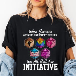 May include: Black t-shirt with the text "When Someone Attacks One Party Member, We All Roll For Initiative." The shirt features colorful geometric dice designs with various symbols. The shirt is worn by a person.
