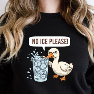 Pode incluir: Camisola preta com um desenho de pato de desenho animado. O pato, com uma expressão zangada, segura um copo de cubos de gelo. Um balão de fala acima do pato diz "NO ICE PLEASE!". Um design humorístico.