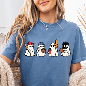 May include: A blue short-sleeved t-shirt featuring a graphic print of four white ghosts dressed for baseball. The ghosts are depicted with a red cap and bat, a blue cap and ball, an orange foam finger and drink, and a black catcher's mask and glove.