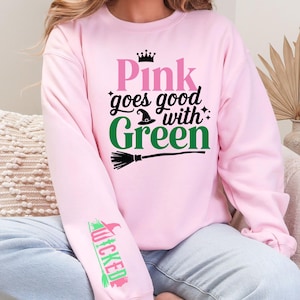 May include: A light pink sweatshirt with the phrase "Pink goes good with Green" in pink and green lettering, a witch hat, and a broom graphic. The sleeve features the word "Wicked" in green and pink. The sweatshirt is a casual style.