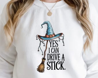 May include: White sweatshirt with a graphic of a blue witch's hat and a brown broom. The text "YES I CAN DRIVE A STICK" is printed in black. A fun, Halloween-themed design. The sweatshirt is a comfortable, casual garment.