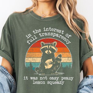 May include: Olive green t-shirt featuring a raccoon illustration within a retro sunset design. The text reads "in the interest of full transparency" and "it was not easy peazy lemon squeezy".