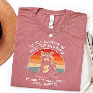 May include: A dusty rose t-shirt with a raccoon graphic inside a sunset-colored circle. The text reads "in the interest of full transparency" and "it was not easy peazy lemon squeezy". A gold necklace and a brown hat are also visible.
