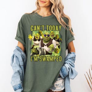 May include: Olive green t-shirt featuring a graphic of Shrek and the text "CAN'T TODAY I'M SWAMPED". The graphic includes Shrek and other characters from the movie. The shirt has short sleeves and a relaxed fit.