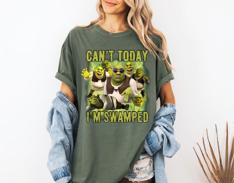 May include: Olive green t-shirt featuring a graphic of Shrek and the text "CAN'T TODAY I'M SWAMPED". The graphic includes Shrek and other characters from the movie. The shirt has short sleeves and a relaxed fit.