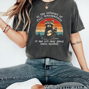 May include: Dark gray t-shirt featuring a raccoon illustration within a sunset design. The text reads "in the interest of full transparency, it was not easy peazy lemon squeazy." The shirt is paired with blue jeans and white shoes.