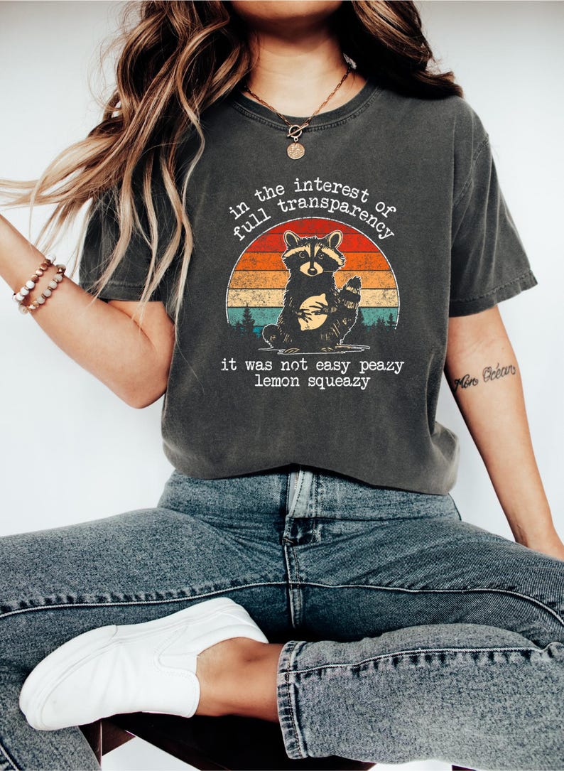 May include: Dark gray t-shirt featuring a raccoon illustration within a sunset design. The text reads "in the interest of full transparency, it was not easy peazy lemon squeazy." The shirt is paired with blue jeans and white shoes.