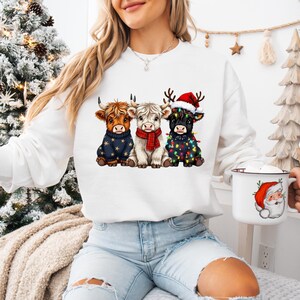 May include: White sweatshirt featuring three Highland cows in festive attire. One cow wears a navy sweater with gold stars, another a red scarf, and the third a Santa hat and Christmas lights. A white mug with a Santa Claus design is also visible.