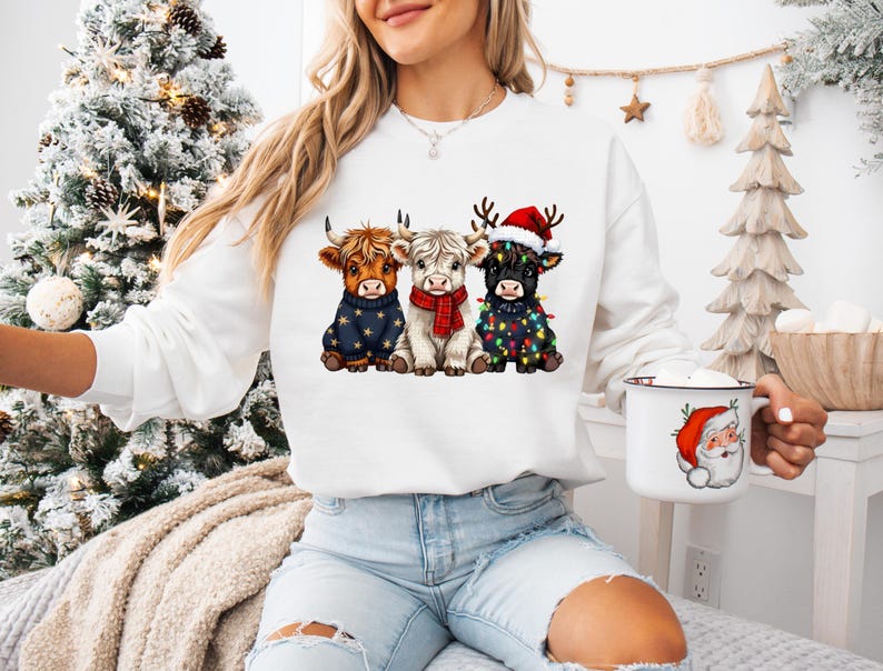 May include: White sweatshirt featuring three Highland cows in festive attire. One cow wears a navy sweater with gold stars, another a red scarf, and the third a Santa hat and Christmas lights. A white mug with a Santa Claus design is also visible.