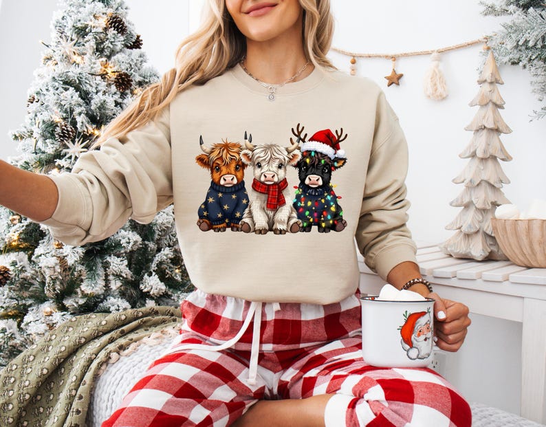 May include: A beige sweatshirt featuring three Highland cows, each adorned with festive holiday attire. One cow wears a blue sweater with stars, another a red scarf, and the third a Santa hat and string lights. A white mug with a Santa Claus graphic is held.