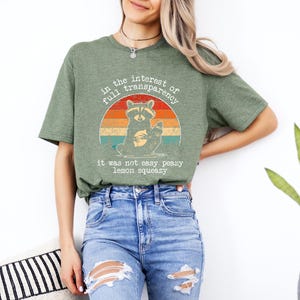 May include: Green t-shirt with a raccoon graphic inside a retro sunset design. The text on the shirt reads "in the interest of full transparency, it was not easy peazy lemon squeezy."