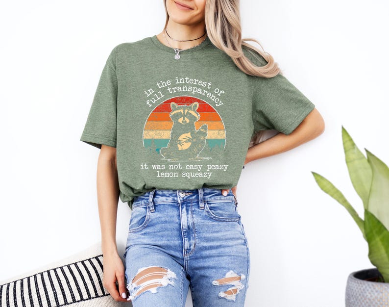 May include: Green t-shirt with a raccoon graphic inside a retro sunset design. The text on the shirt reads "in the interest of full transparency, it was not easy peazy lemon squeezy."