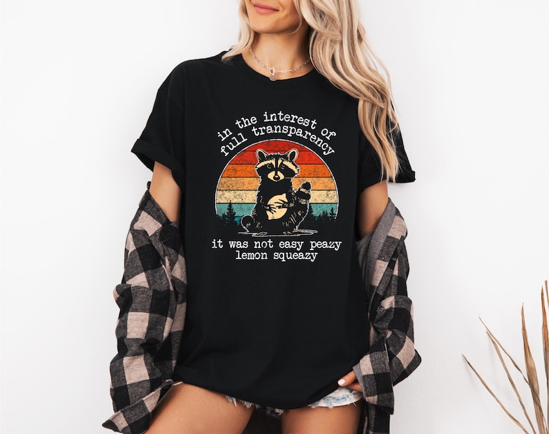 May include: Black t-shirt featuring a raccoon graphic inside a sunset design. The text reads "in the interest of full transparency, it was not easy peazy lemon squeezy". The shirt is a casual wear item.