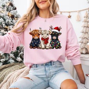 May include: A light pink sweatshirt featuring a graphic of three Highland cows dressed for Christmas. One cow wears a blue sweater, another a red scarf, and the third a Santa hat and string lights. The sweatshirt is worn by a person.