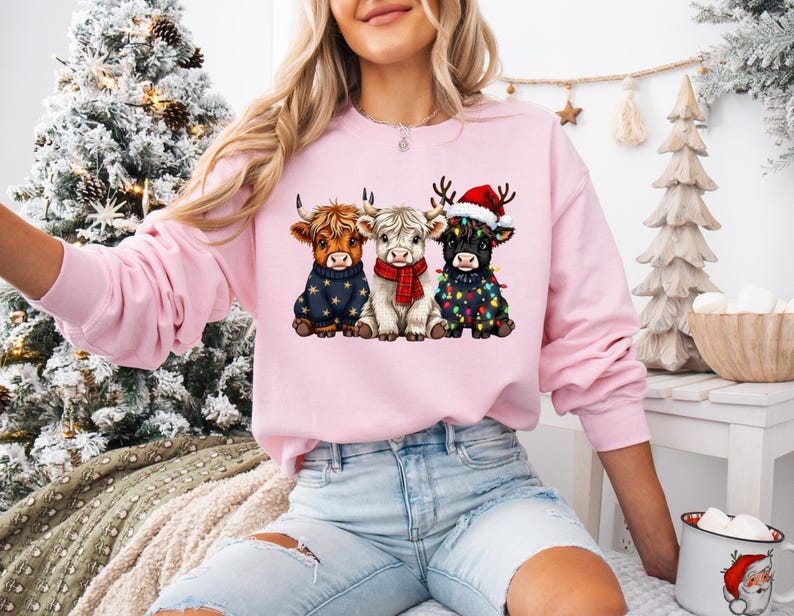 May include: A light pink sweatshirt featuring a graphic of three Highland cows dressed for Christmas. One cow wears a blue sweater, another a red scarf, and the third a Santa hat and string lights. The sweatshirt is worn by a person.