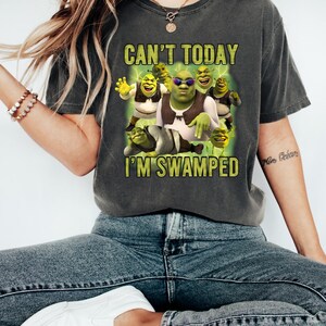 May include: Dark gray t-shirt featuring a graphic of Shrek and other characters with the text "CAN'T TODAY I'M SWAMPED" in yellow. The image shows Shrek wearing sunglasses and a vest. The shirt is worn by a person wearing blue jeans and white shoes.