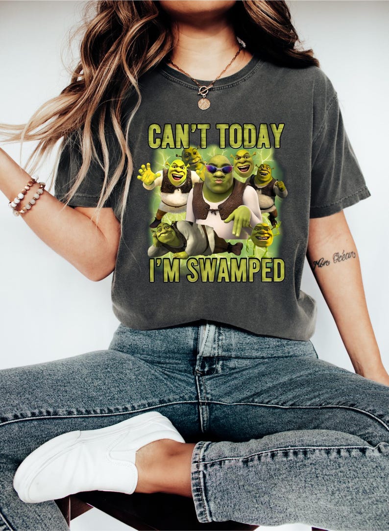May include: Dark gray t-shirt featuring a graphic of Shrek and other characters with the text "CAN'T TODAY I'M SWAMPED" in yellow. The image shows Shrek wearing sunglasses and a vest. The shirt is worn by a person wearing blue jeans and white shoes.