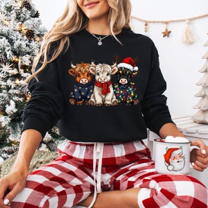 May include: Black sweatshirt featuring three Highland cows in festive attire. One cow wears a Santa hat, another is adorned with Christmas lights, and the third is in a blue sweater. The model is wearing red and white plaid pajama pants and holding a Santa mug.